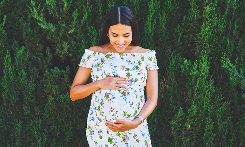 A woman stands in a floral dress, smiling while looking downwards and embracing her round, pregnant belly.