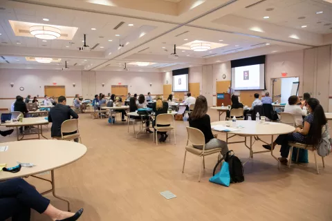 Doordan Institute GME Conference Room, with attendees sitting at round tables and a speaker giving a presentation at a podium at the front of the room.