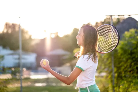 A young girl in the morning summer sun, holding a tennis racket high and preparing to serve.
