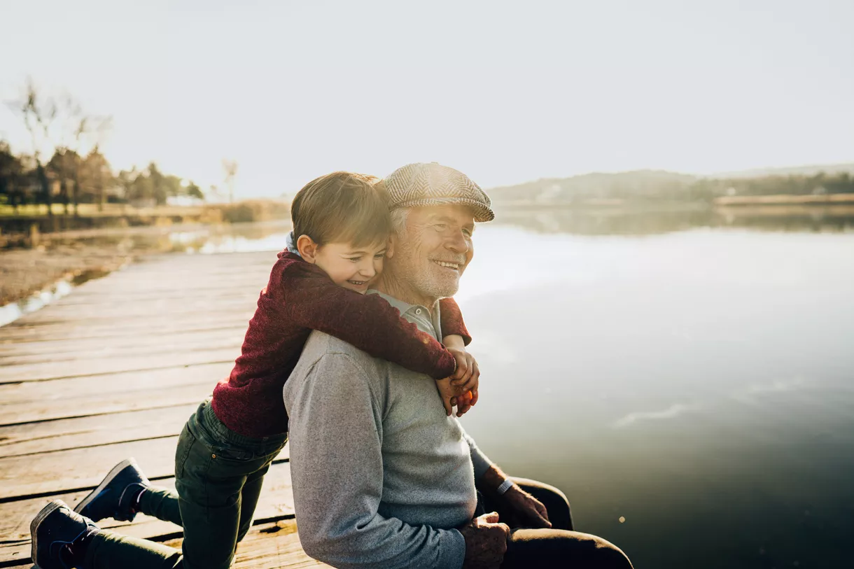 A grandfather sits on a dock, being embraced by his grandson.