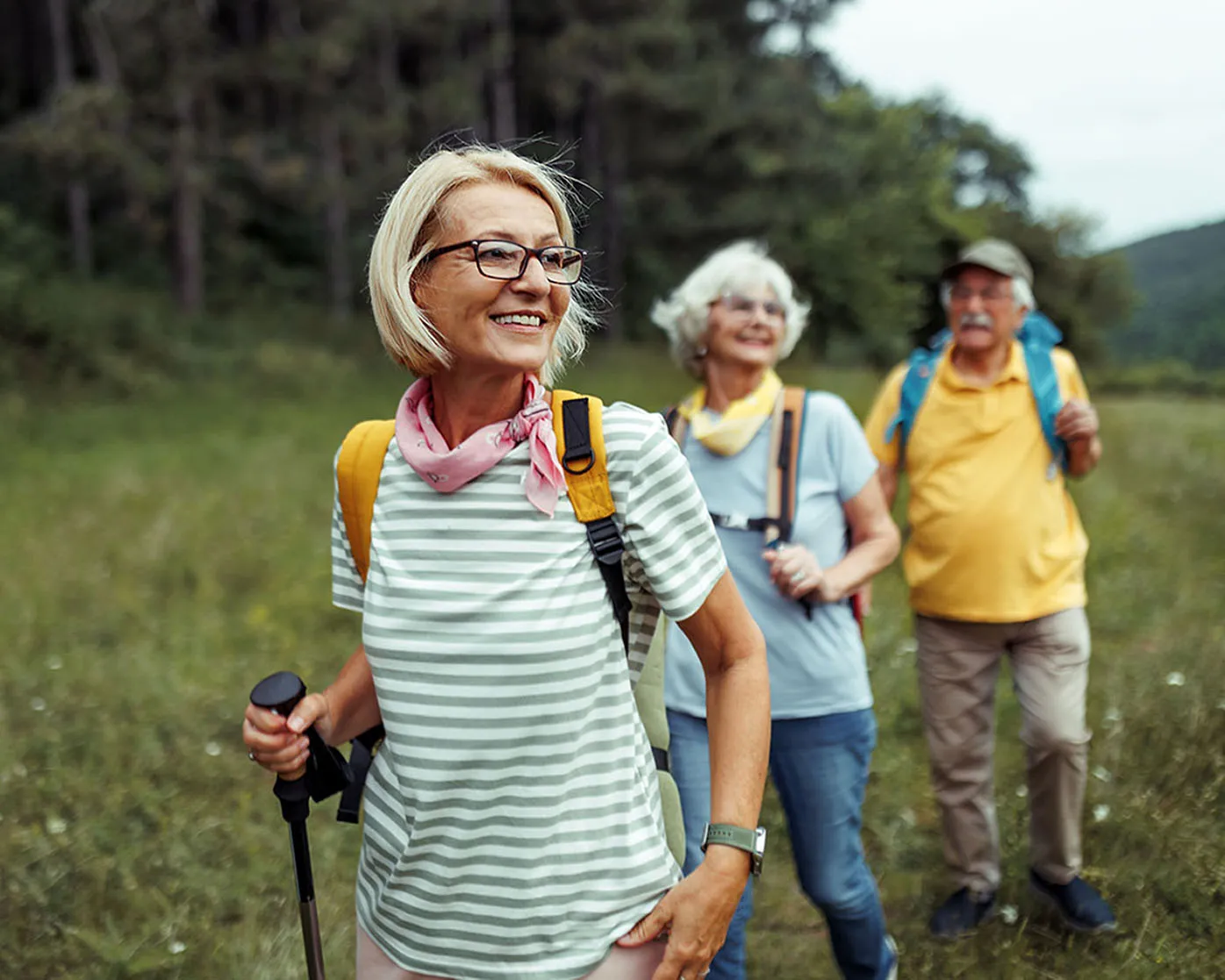 A woman smiling while hiking in an open field with trees nearby. An older couple that look to be her parents are not far behind. 